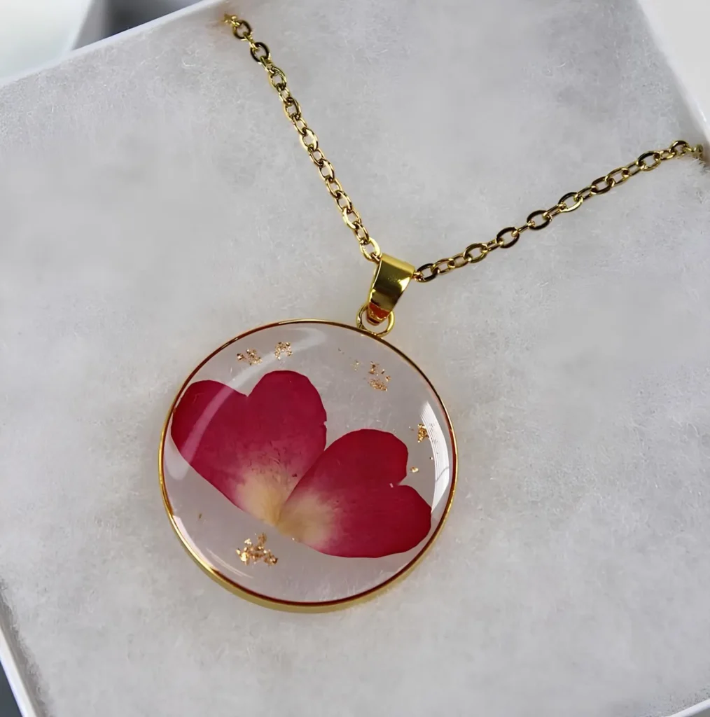 A round resin flower pendant necklace featuring preserved red petals and delicate gold flakes inside a clear dome, set in a gold bezel and suspended on a gold chain, displayed on a soft white background.
