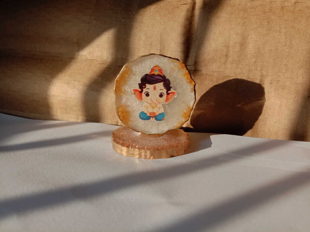 Round resin art coaster featuring a cute Lord Ganesha illustration with gold edges, placed on a matching stand in warm natural light. Festive Décor Ideas Using Resin Art