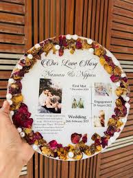 Resin Art Choices A personalized resin memory frame decorated with dried flowers around the border, featuring printed photos and event details, held in hand against a wooden background. Handmade Custom vs Ready Resin Pieces: A Complete Comparison